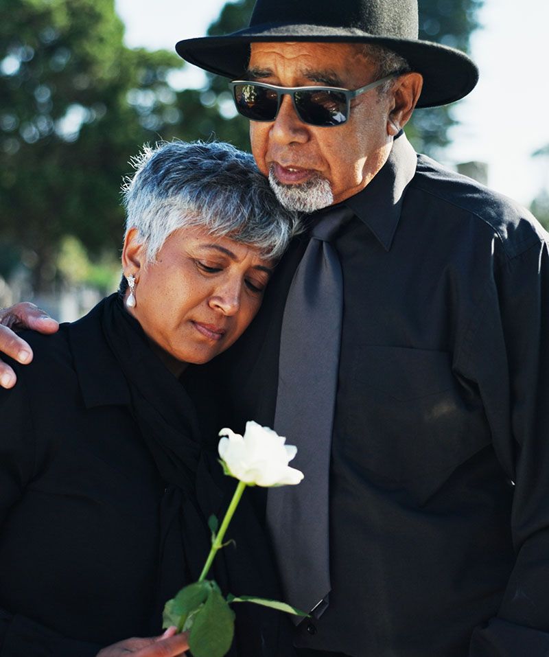 Two people in black clothing embrace at a memorial, holding a white flower.