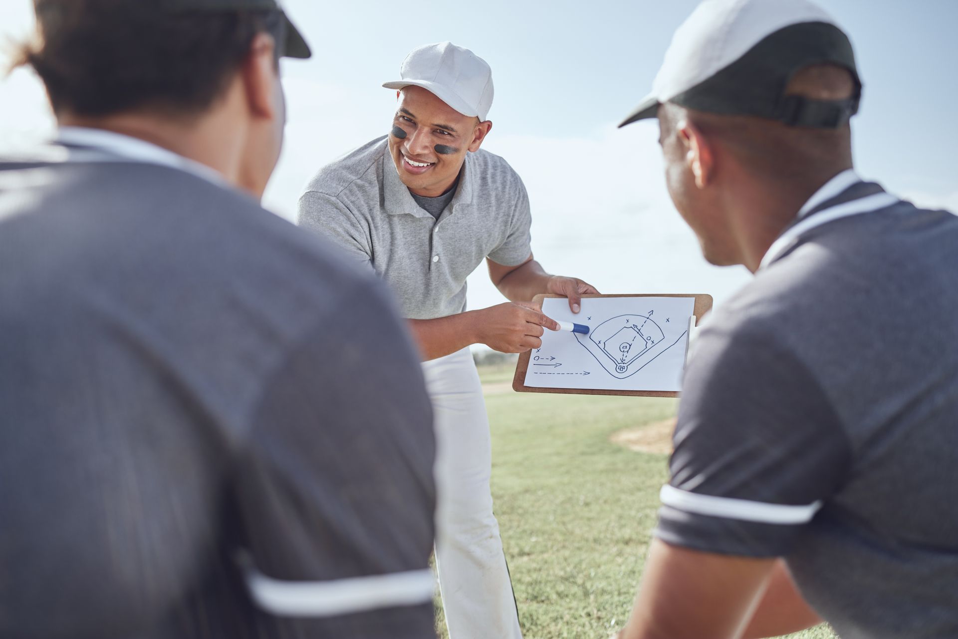 Coach explaining a play on a clipboard to two players on a green field. All wear caps and polos.