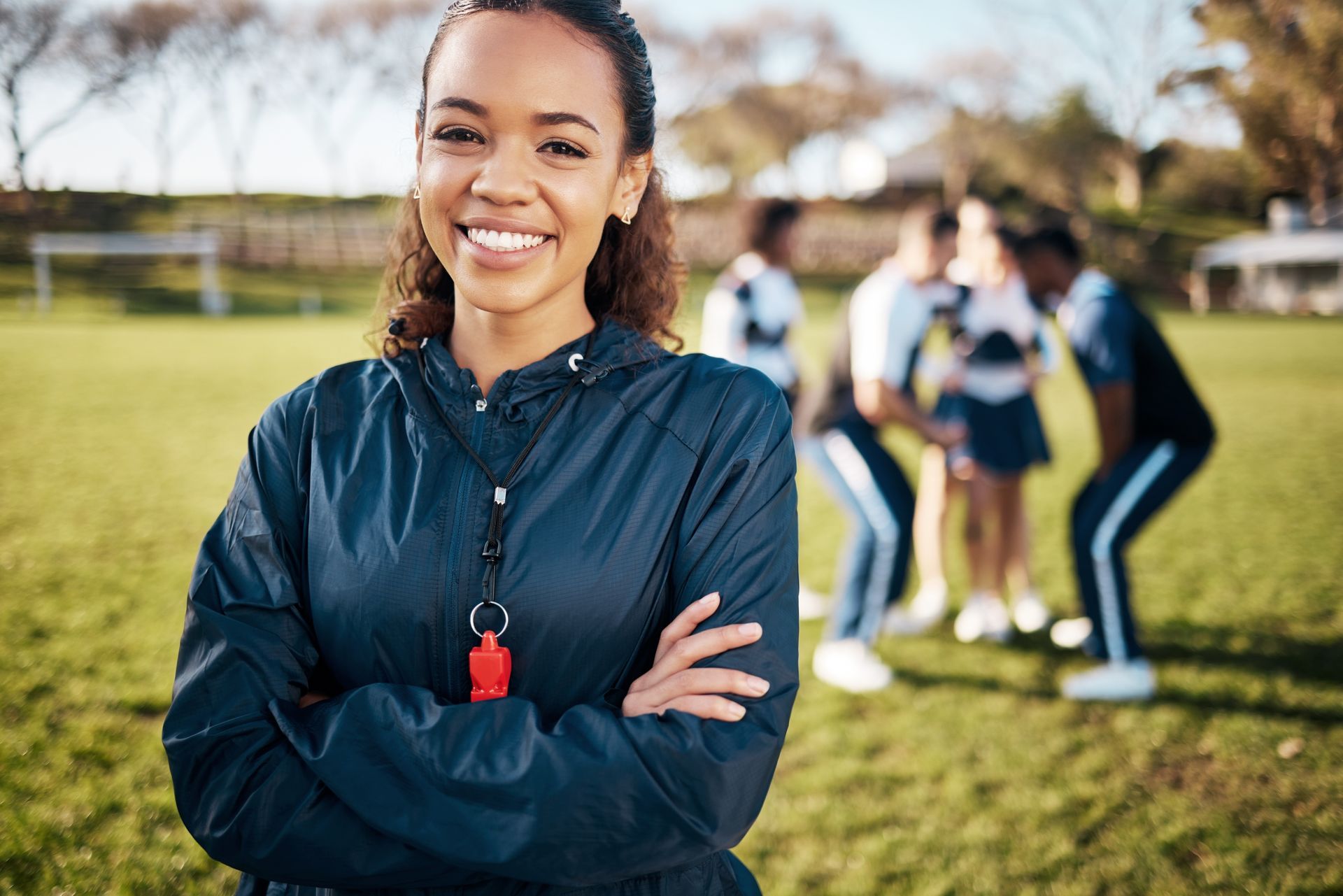 Woman in navy jacket, arms crossed, smiles at camera. Team huddle in the background on a grassy field.