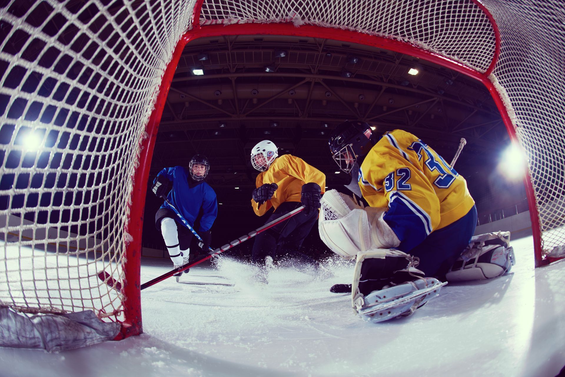 Hockey players in action near net: goalie in white, two attackers in blue and yellow. Ice, net.