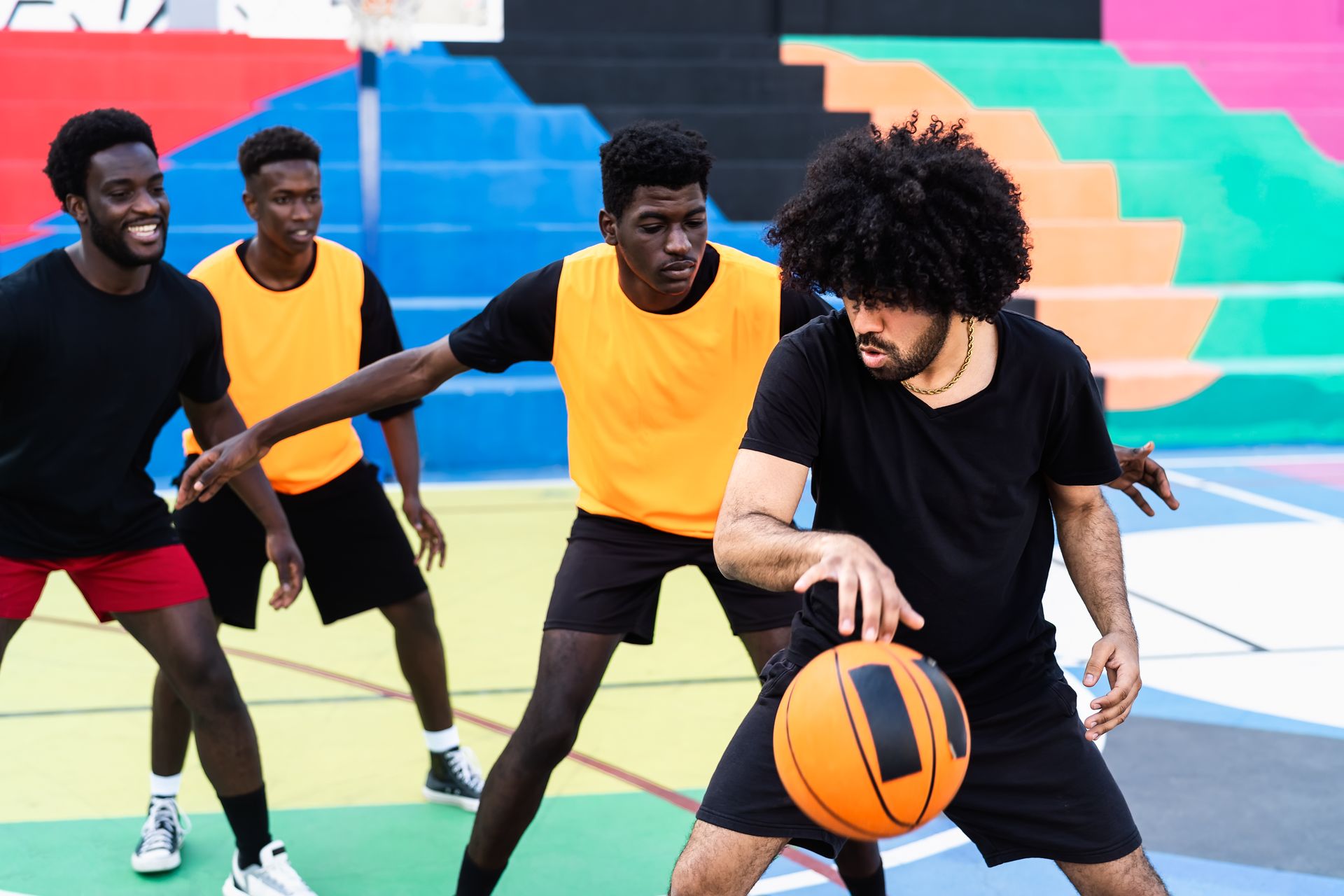 Basketball players on an outdoor court. One dribbles, others defend; colorful background.