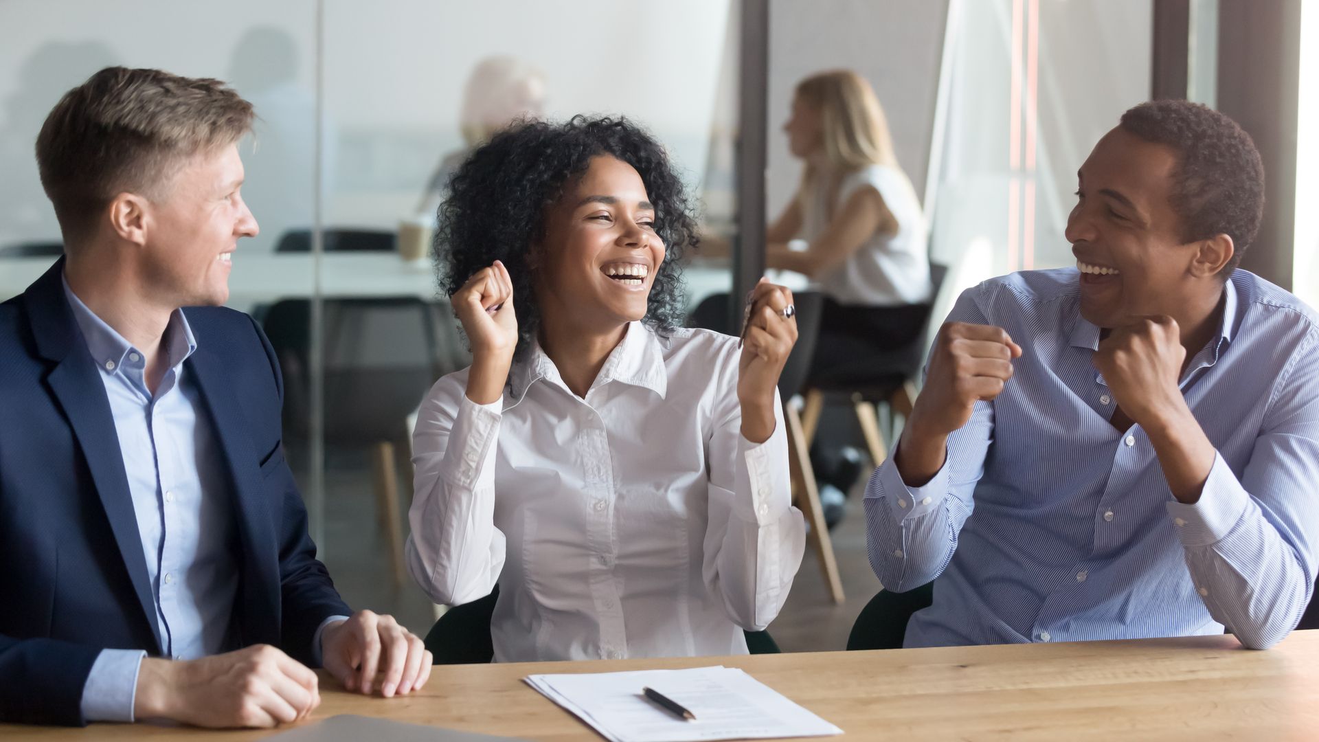 Three coworkers in a bright office celebrate with raised fists and smiles around a table.