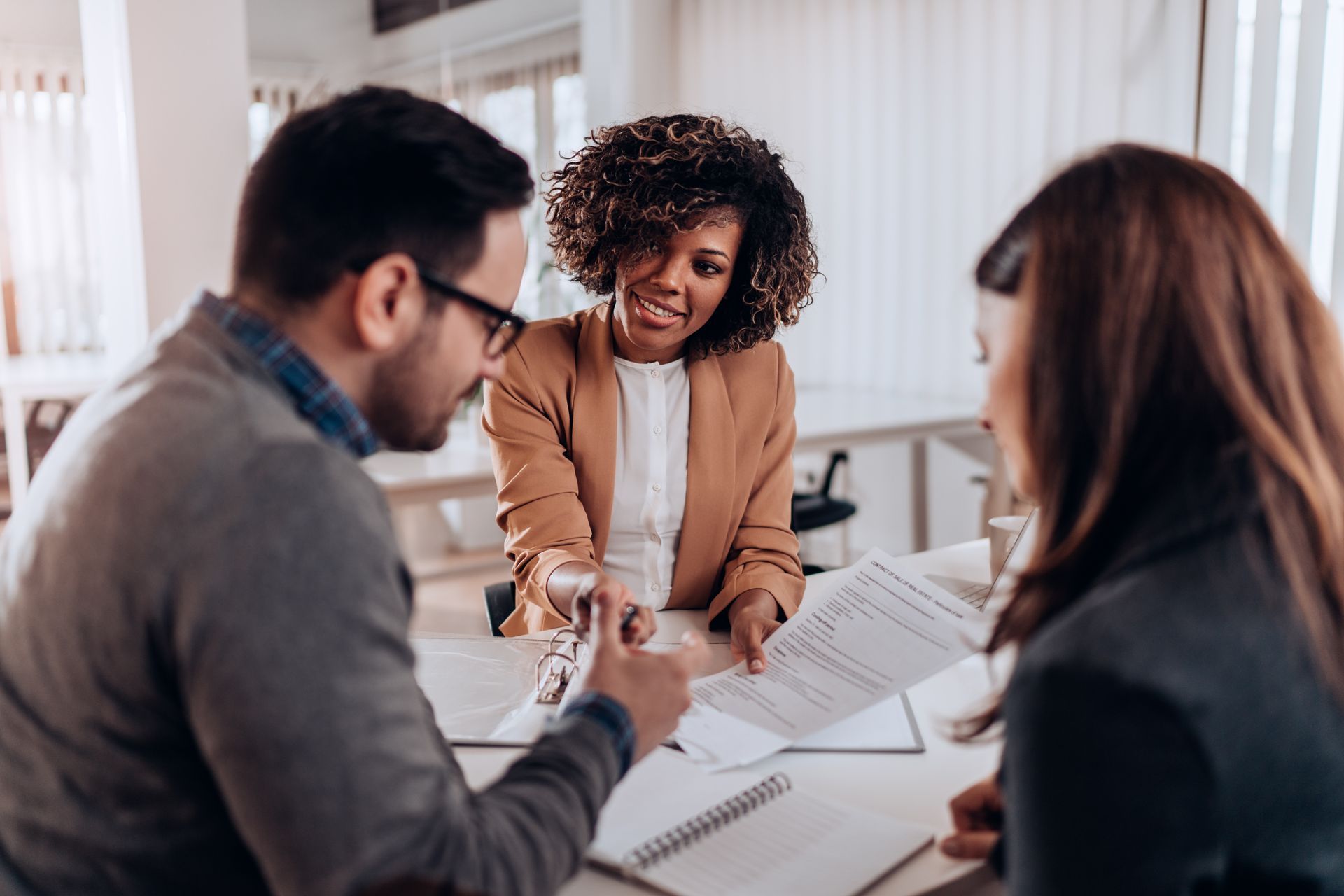 Three people in a bright office discuss documents at a table, with one smiling and pointing at papers.
