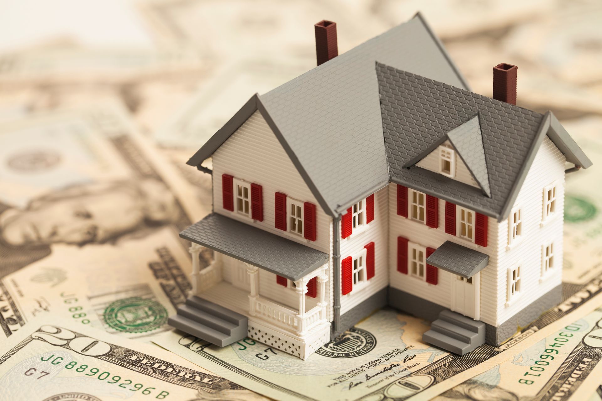 Small white house model with red accents sitting on U.S. dollar bills, symbolizing real estate and money