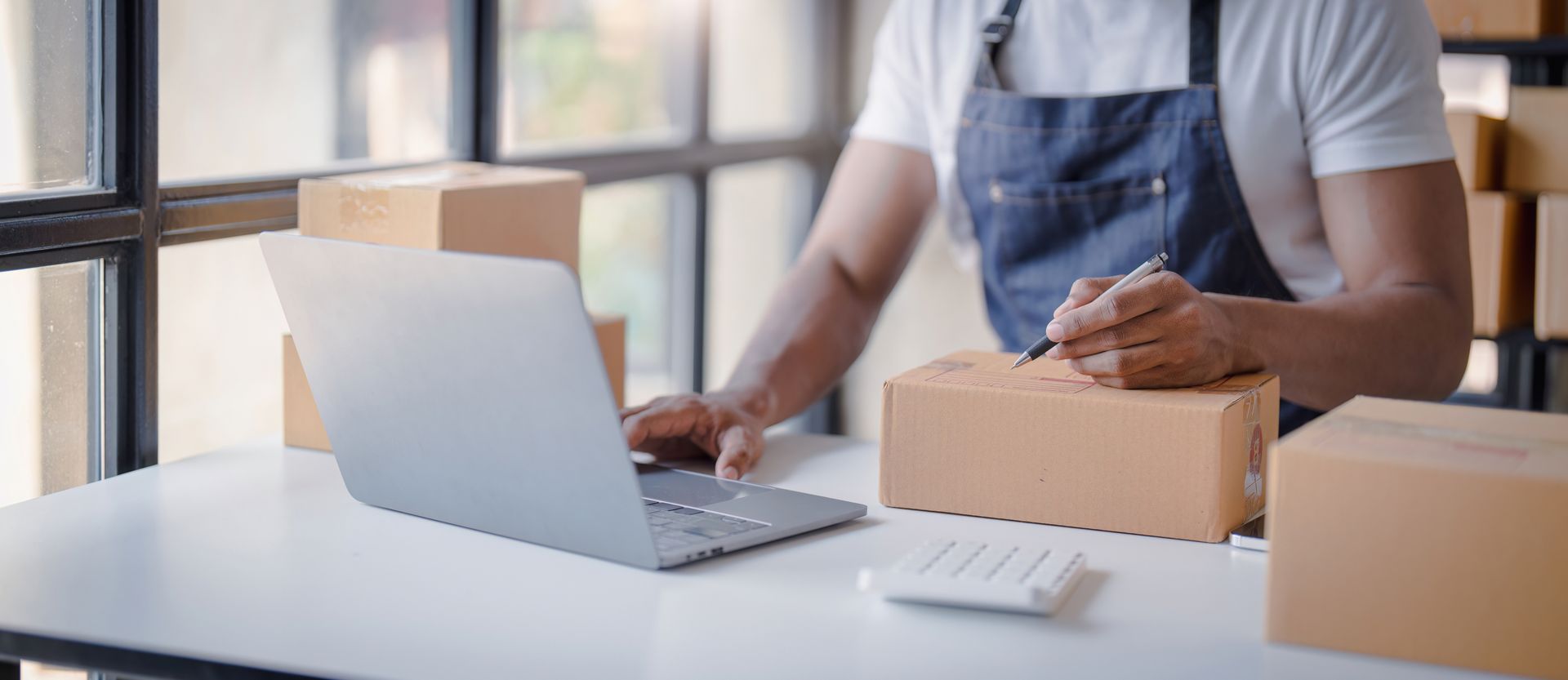 Person packing boxes beside a laptop at a bright desk in an office or home workspace