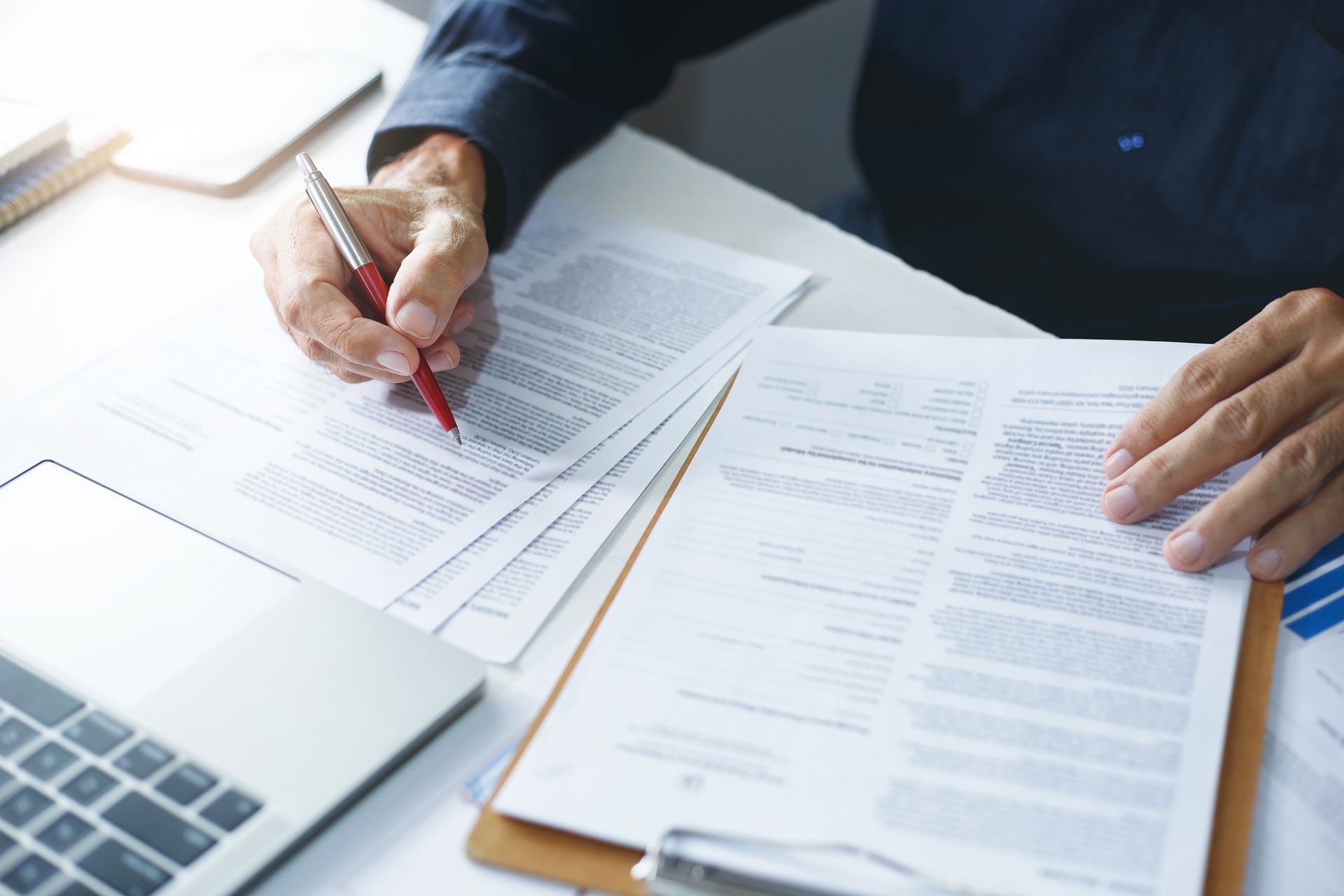 Hands reviewing paperwork beside a laptop, with one person marking documents with a red pen.