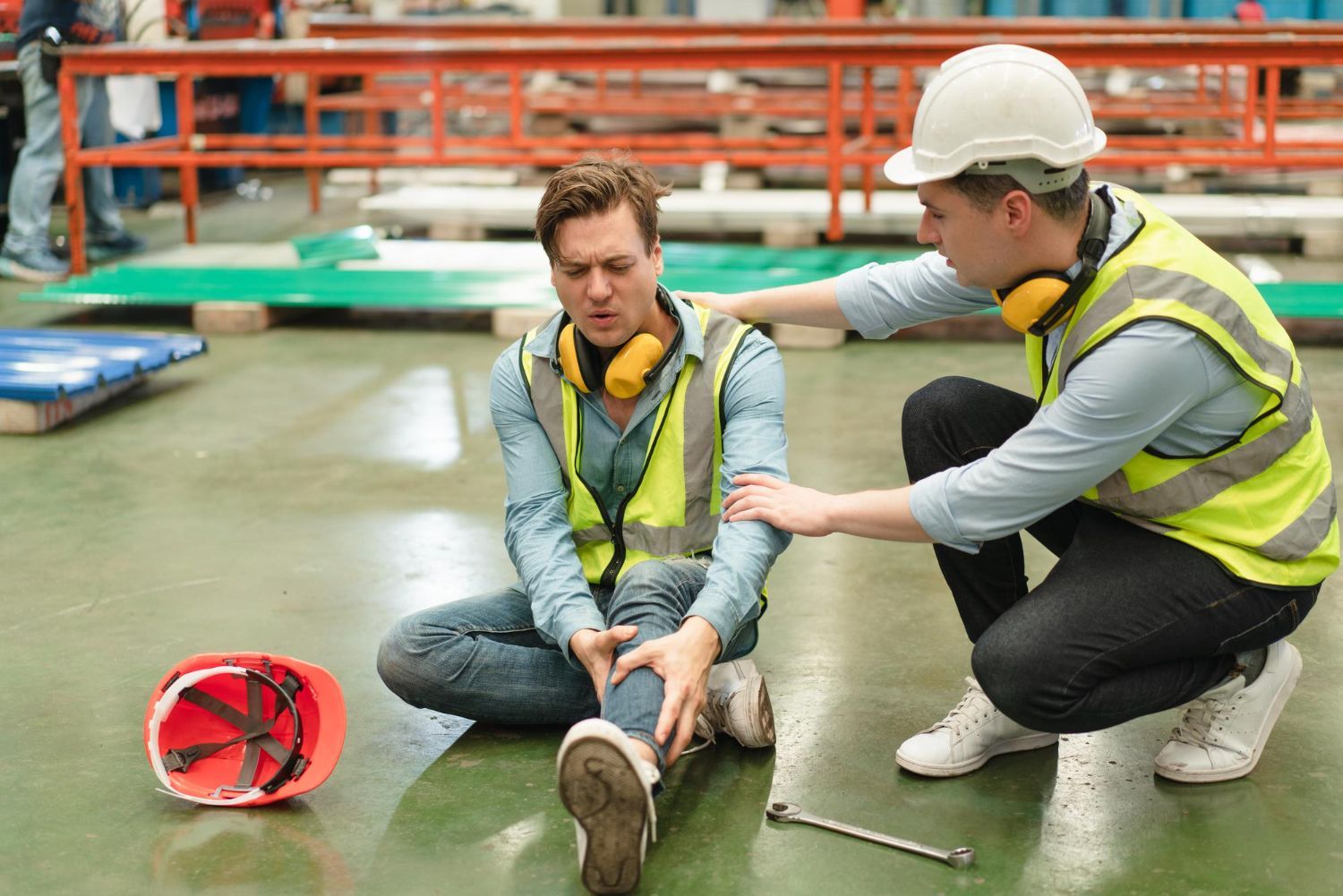 Man injured on factory floor, supported by another worker. Hard hat and wrench nearby.