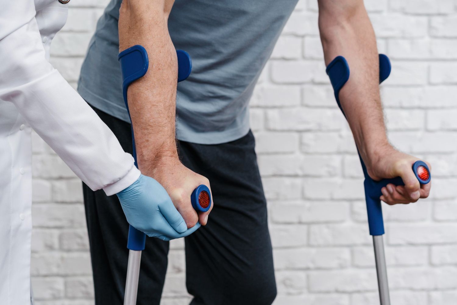Person using crutches, assisted by a person wearing a medical coat and gloves, against a white brick wall.