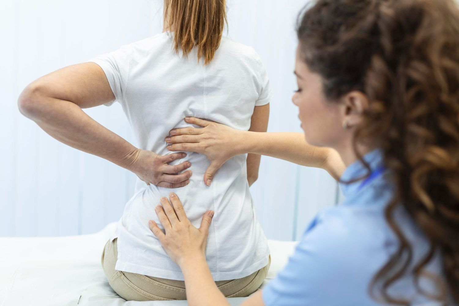 Medical professional examining a person's lower back for pain, hands on the patient's spine, light setting.