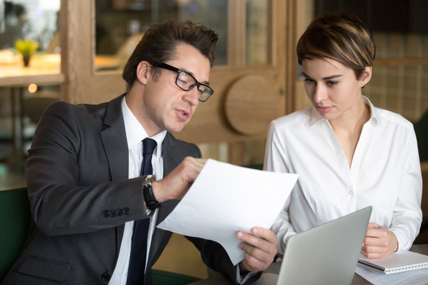 Man in suit points to paper; woman in white shirt looks at laptop. Indoors, business discussion.