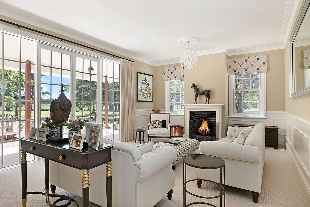 Bright living room with white sofas, fireplace, chandelier, and large windows overlooking a garden.