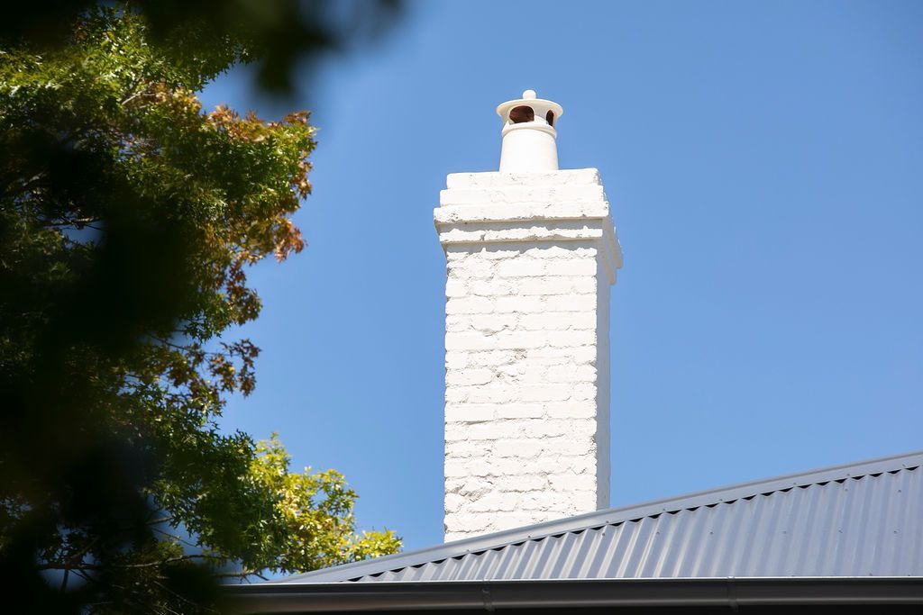 White chimney above a gray roof against a blue sky, framed by green tree leaves