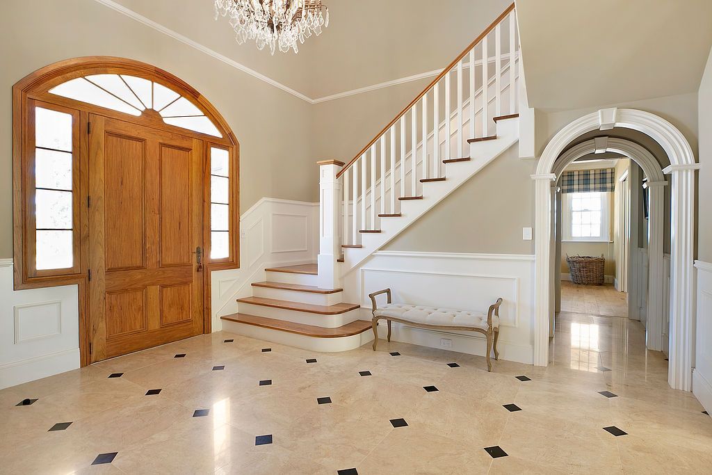 Bright foyer with staircase, arched wooden door, chandelier, and black-and-white tile floor