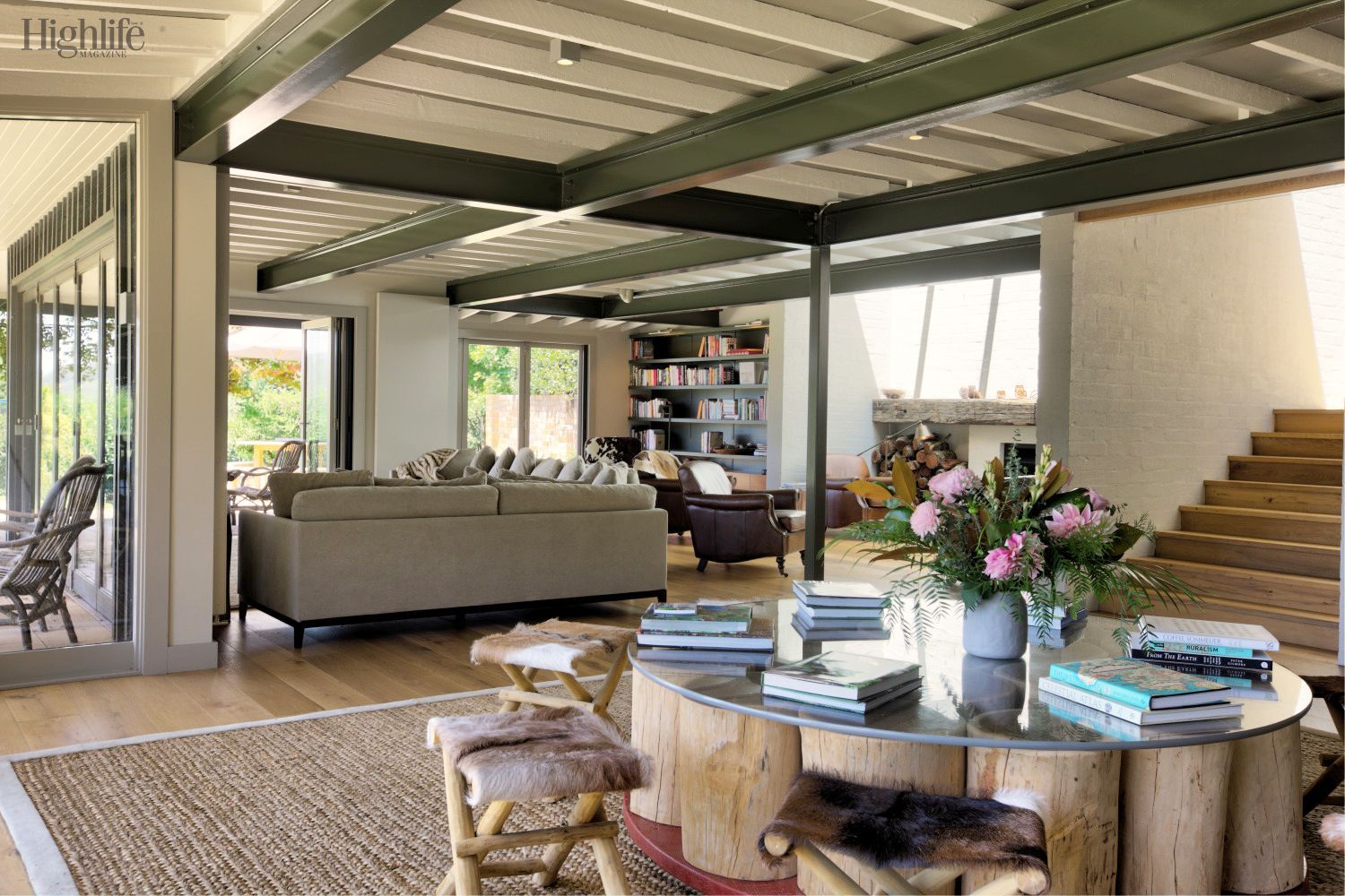 Modern open-plan living room with sofas, coffee table, bookshelves, and large windows overlooking greenery