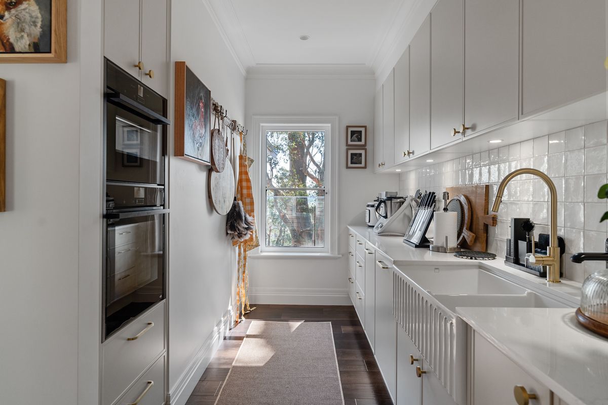 Bright white galley kitchen with built-in ovens, gold faucet, and a window at the end.