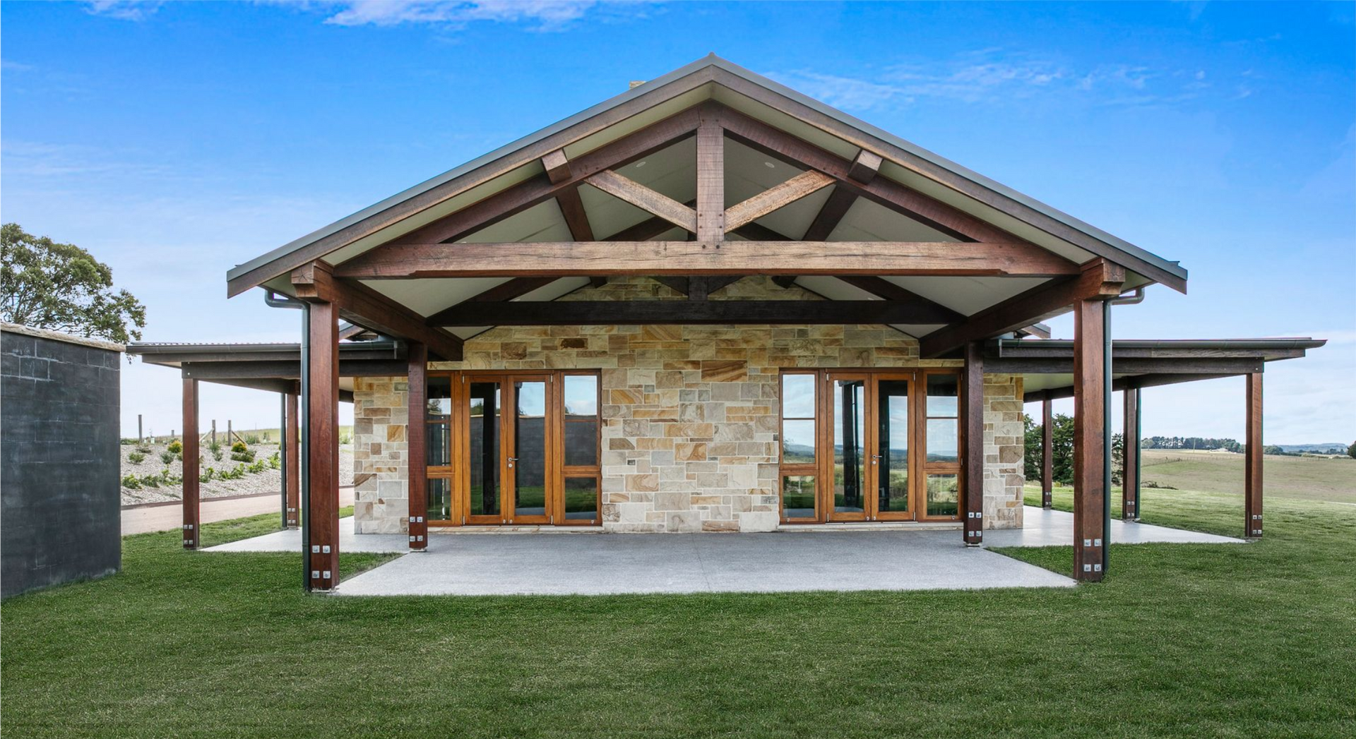 Modern stone house with a wooden porch, exposed beams, and a green lawn under a clear sky