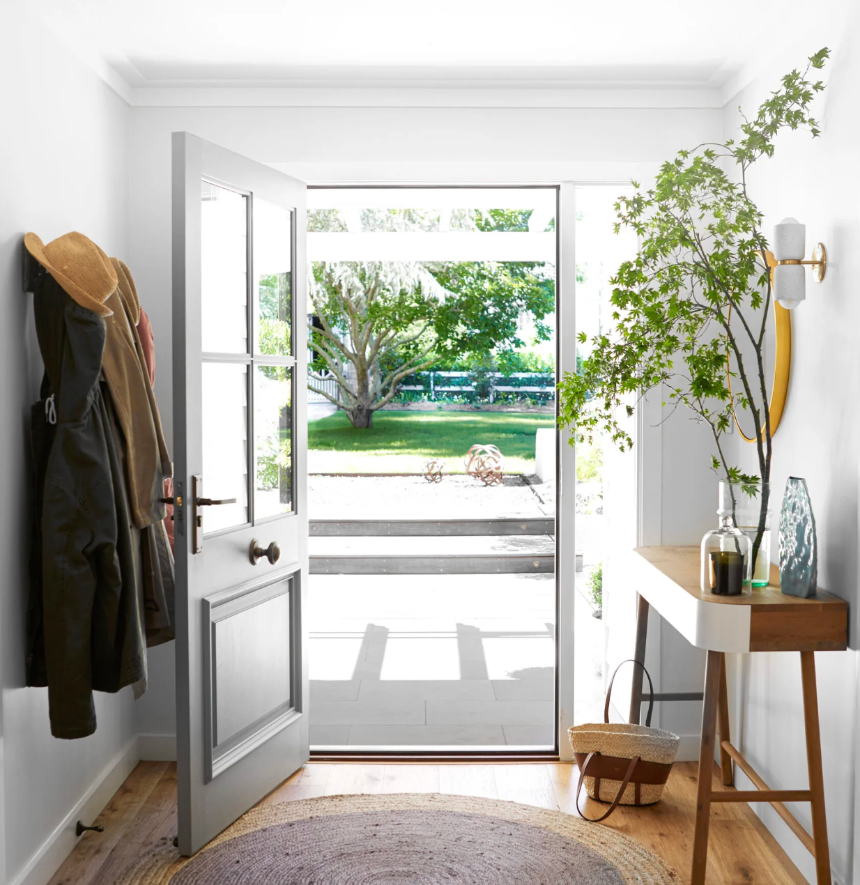 Bright entryway with open front door, coat rack, and small table, looking out to a sunny garden.