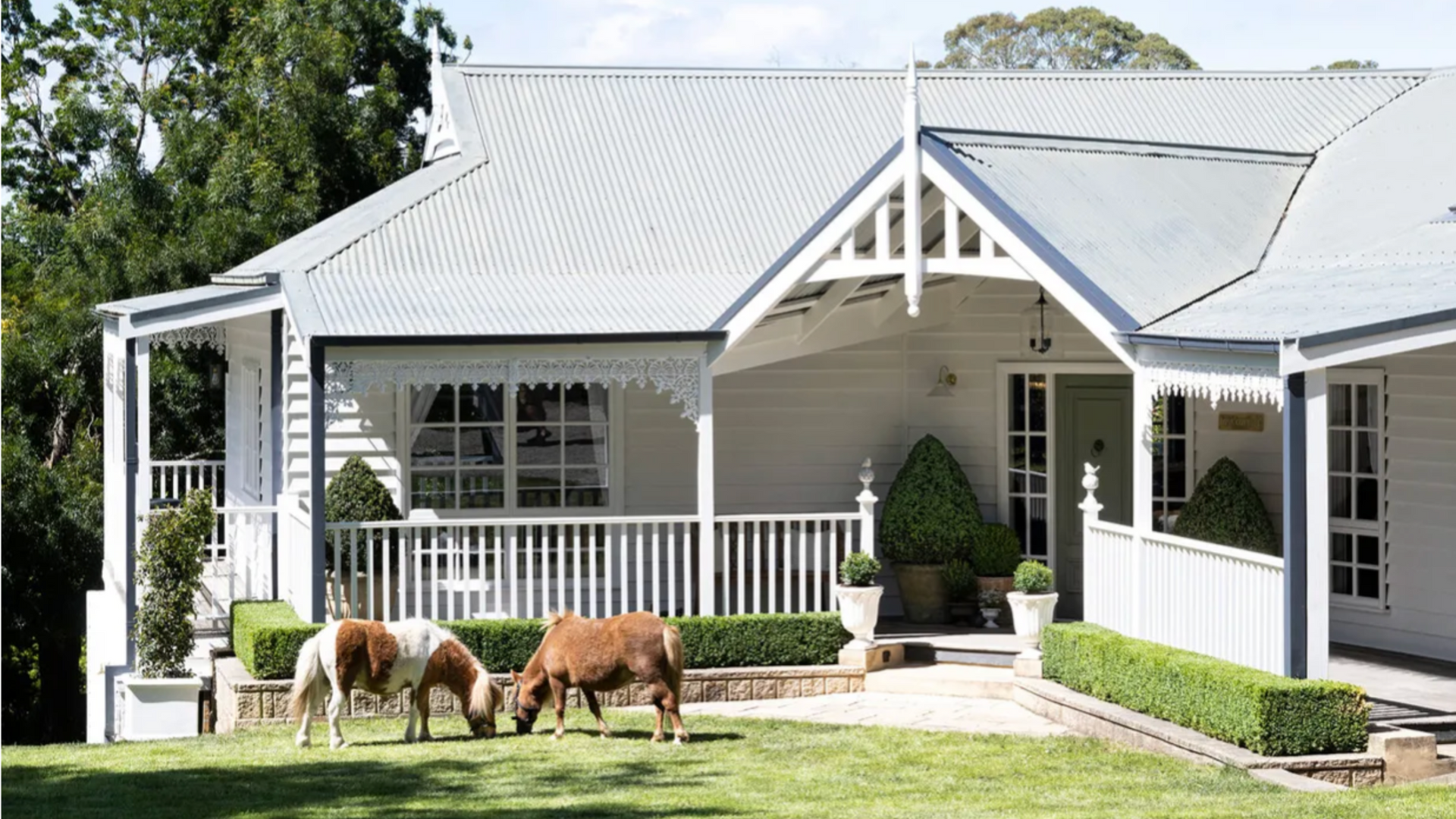 White cottage with porch and two grazing horses on the front lawn