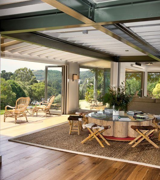 Modern open patio with glass walls, wooden floor, woven chairs, round table, and greenery outside