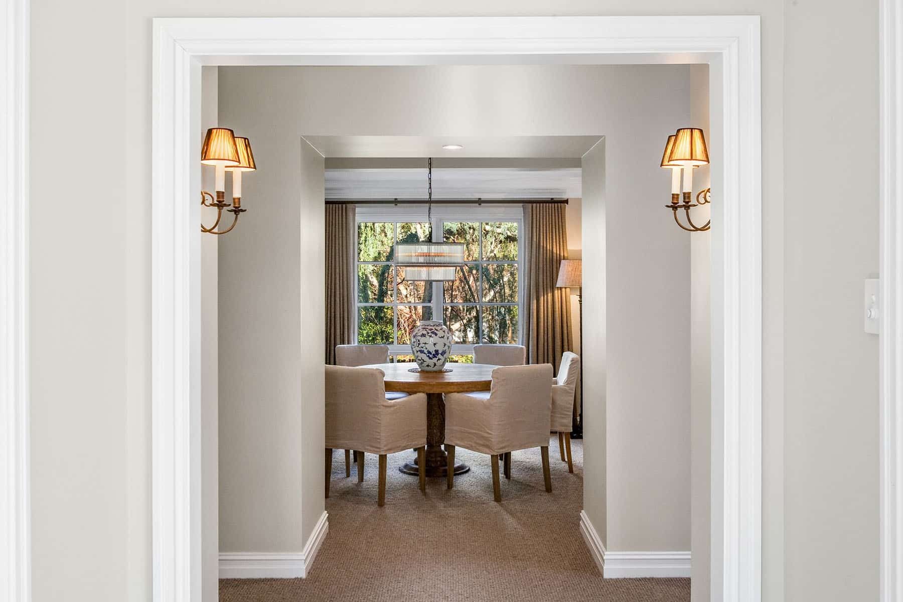 Elegant hallway leading to a sunlit dining room with upholstered chairs and wall sconces