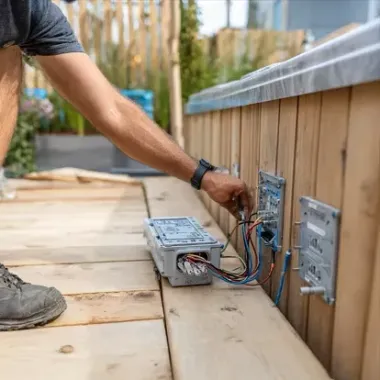 Person wiring an outdoor electrical box beside a wooden deck railing.
