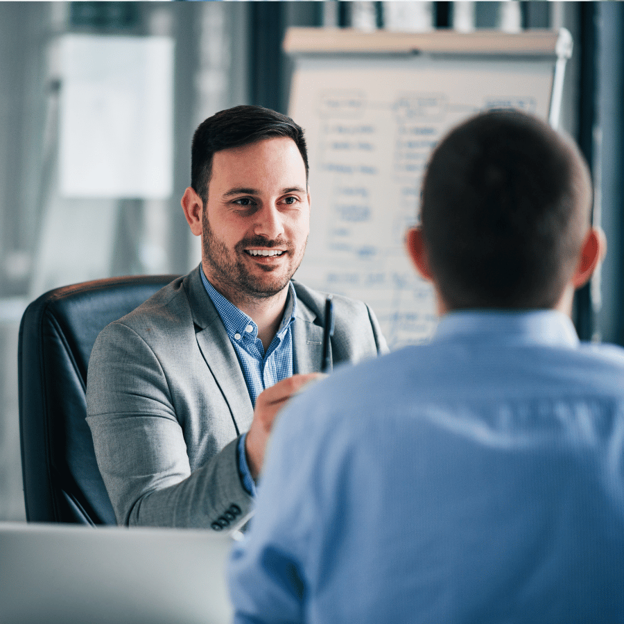 Two people in a business meeting, one speaking across a desk in a bright office