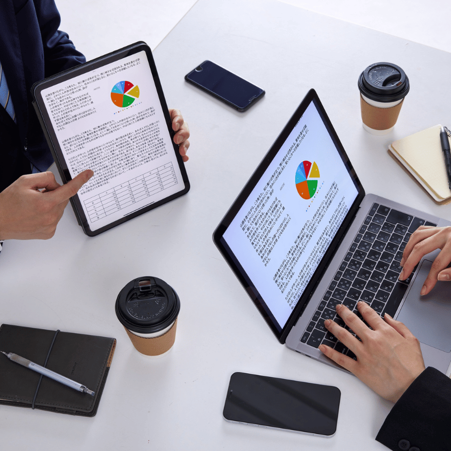Two people review charts on a tablet and laptop at a desk with coffee cups and phones.