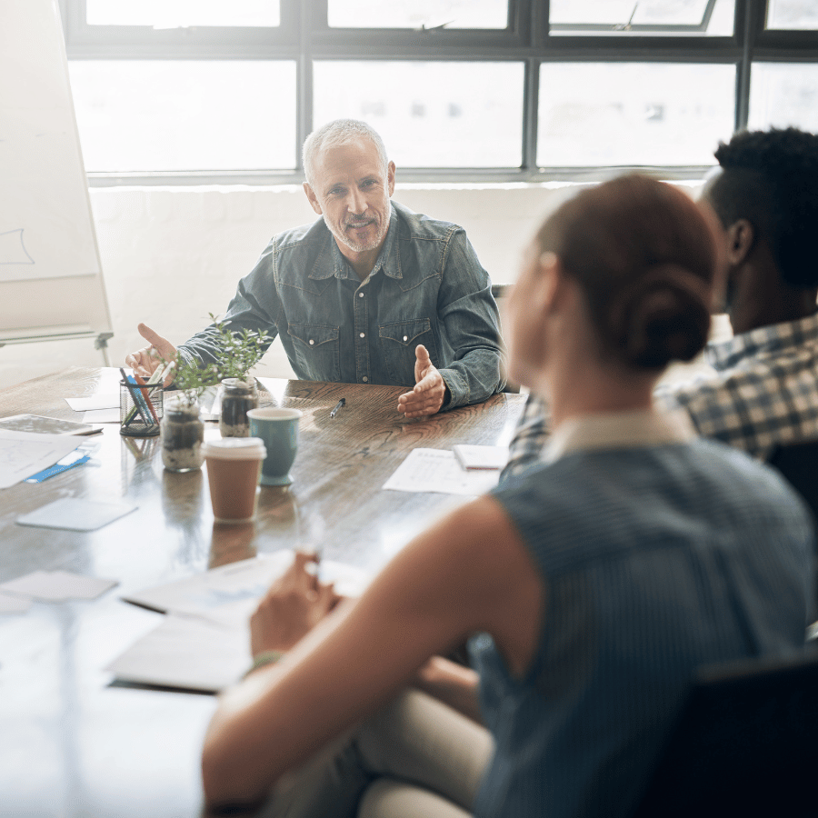 Meeting in a bright office with three people around a table, discussing papers and coffee cups