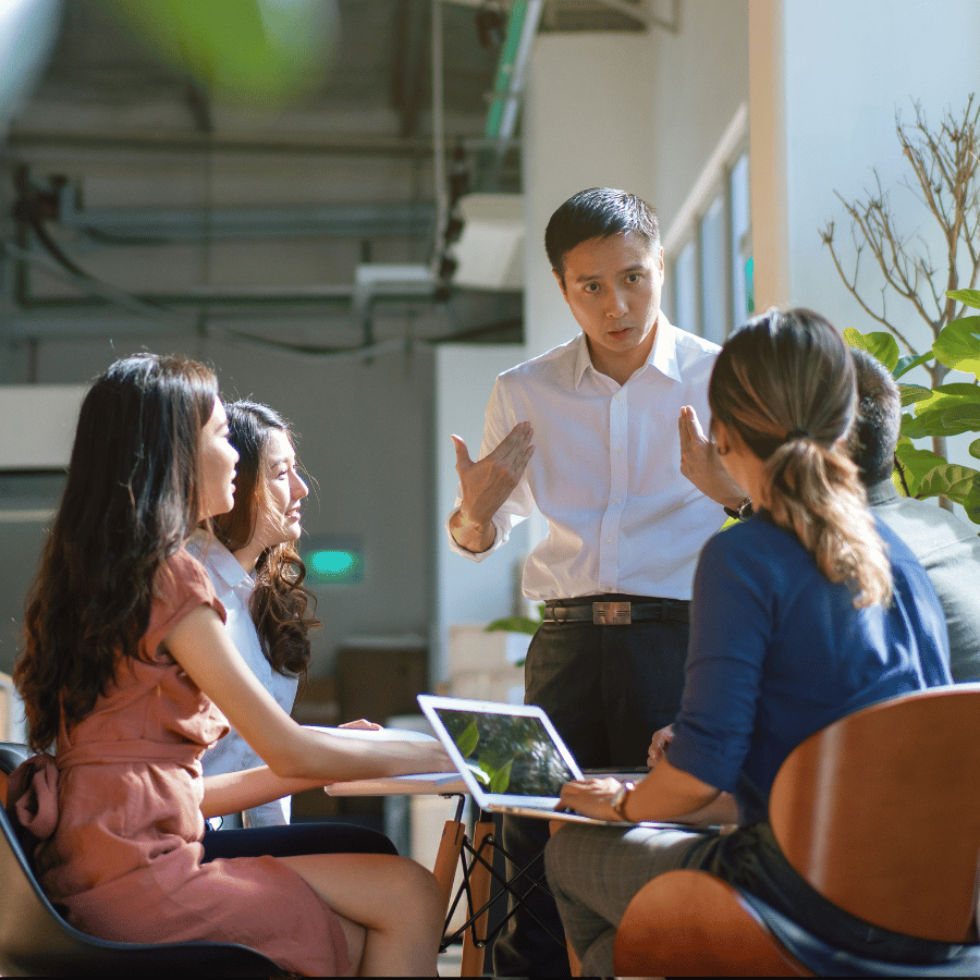 Team meeting in a bright office, with one person presenting to four seated colleagues around a laptop.