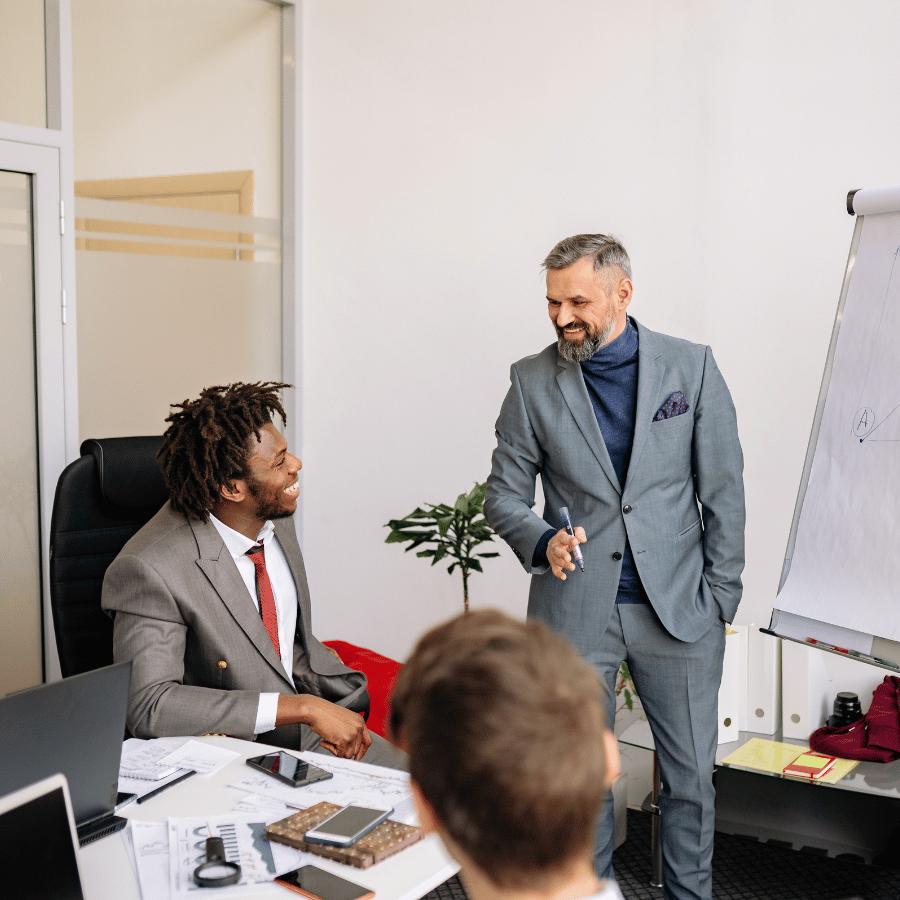 Business meeting in an office, with a man presenting beside a whiteboard and two colleagues seated nearby.