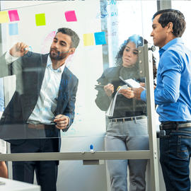 Three coworkers brainstorming at a glass wall with sticky notes and markers in an office.