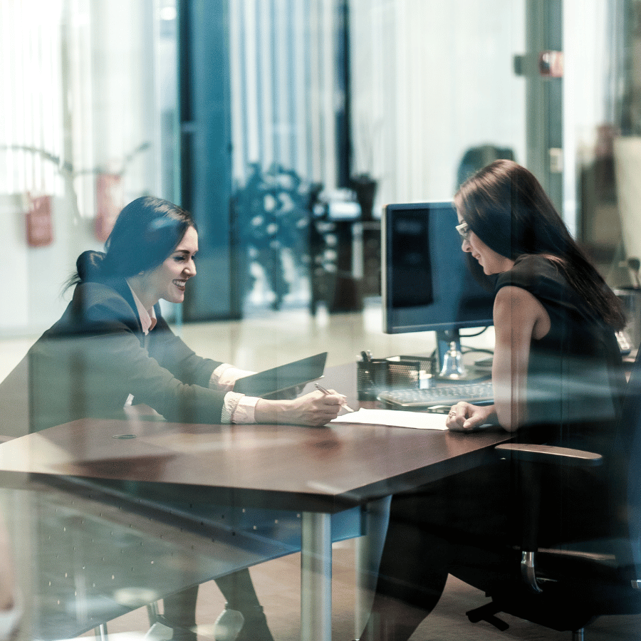 Two people talk across a conference table in a glass-walled office.