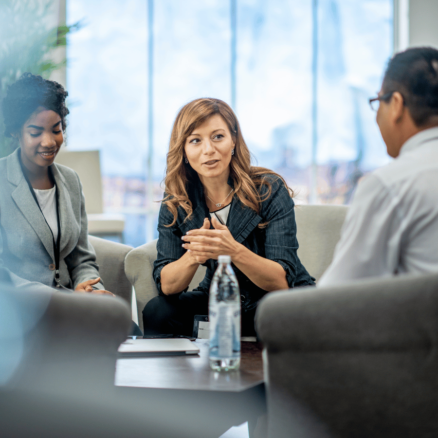 Three people in a meeting, seated around a table in a bright office, speaking attentively.