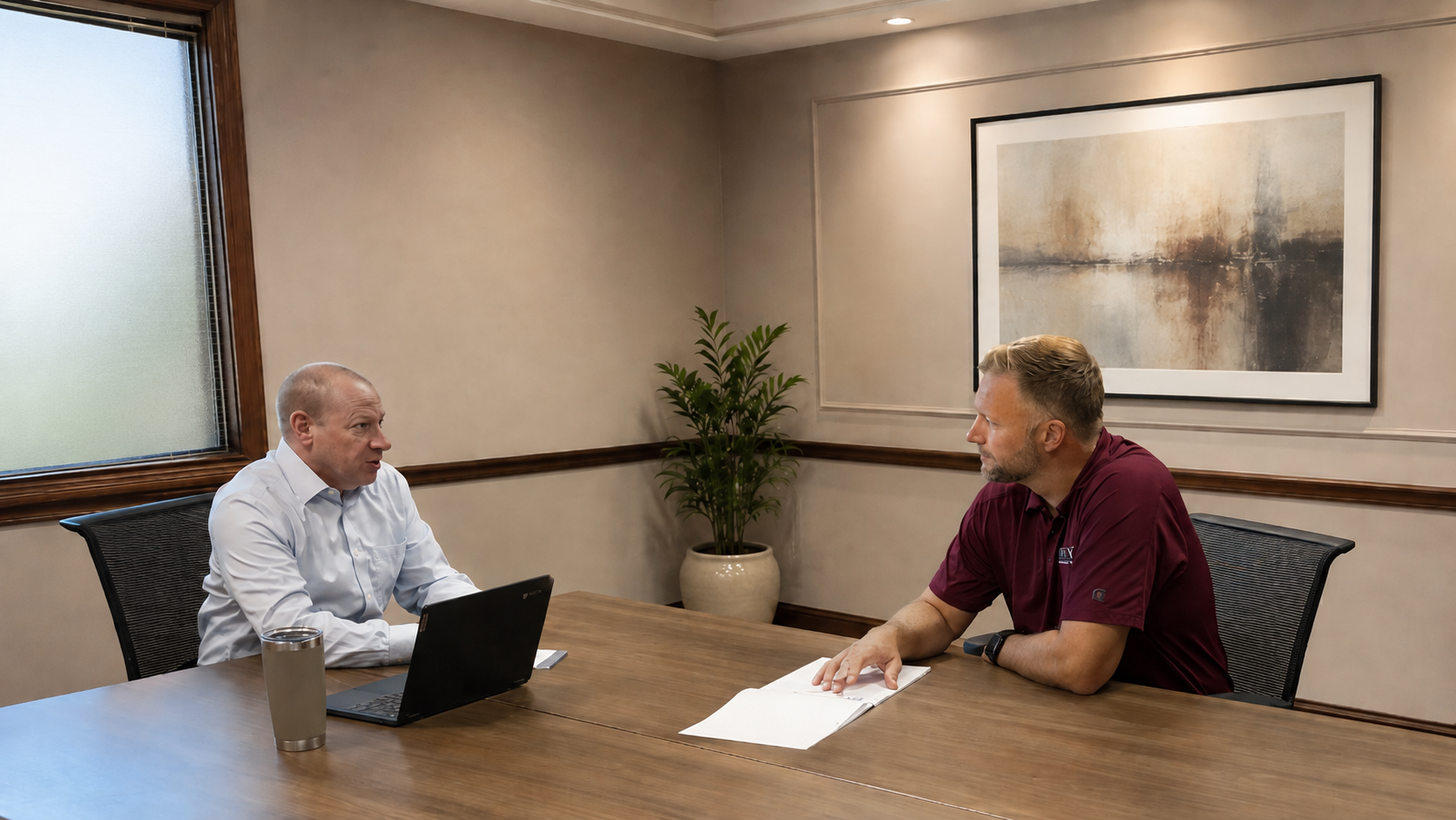 Two people meeting across a conference table in a small office, with a laptop, papers, and wall art.
