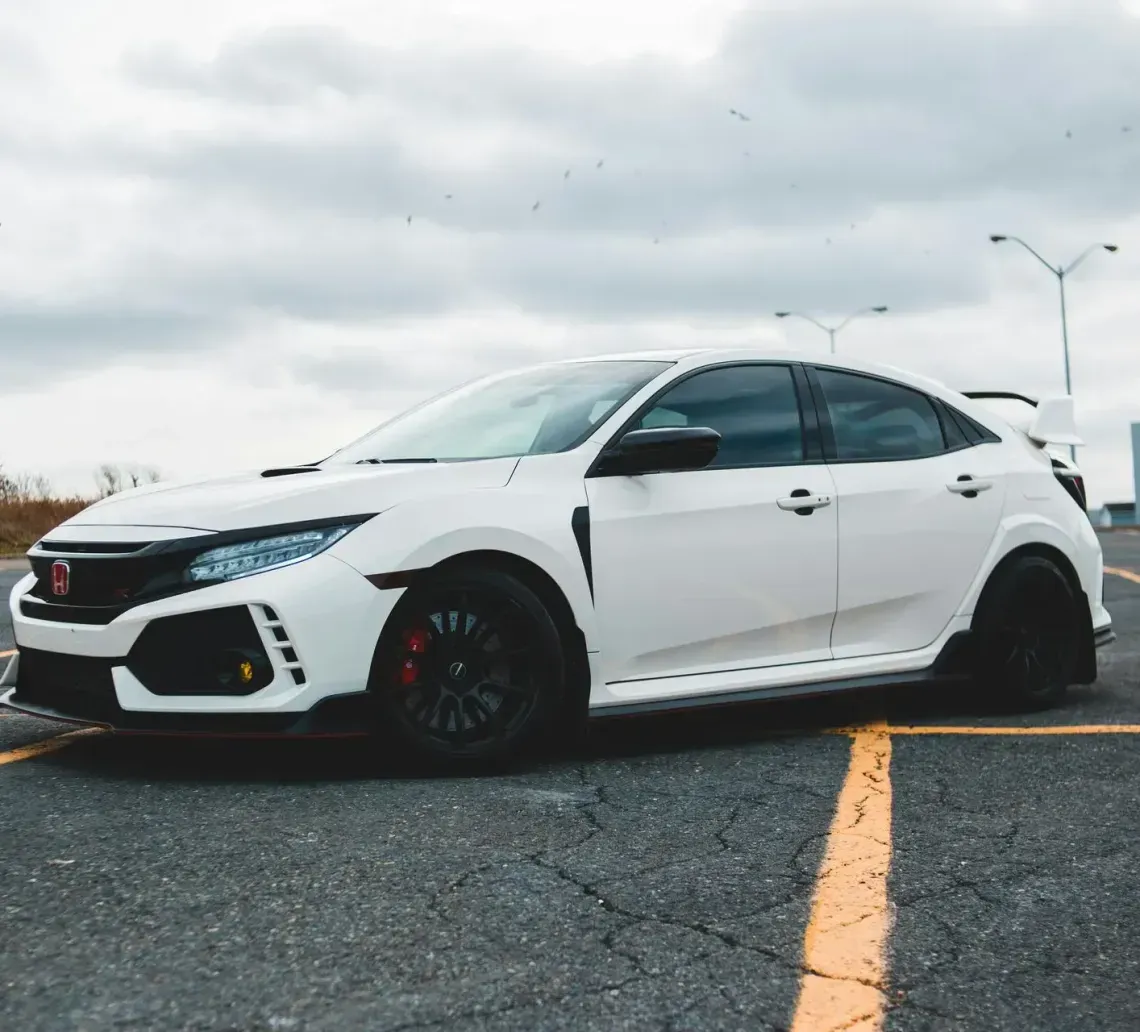 White Honda Civic Type R parked on asphalt, cloudy sky overhead. Black wheels, red brake calipers.