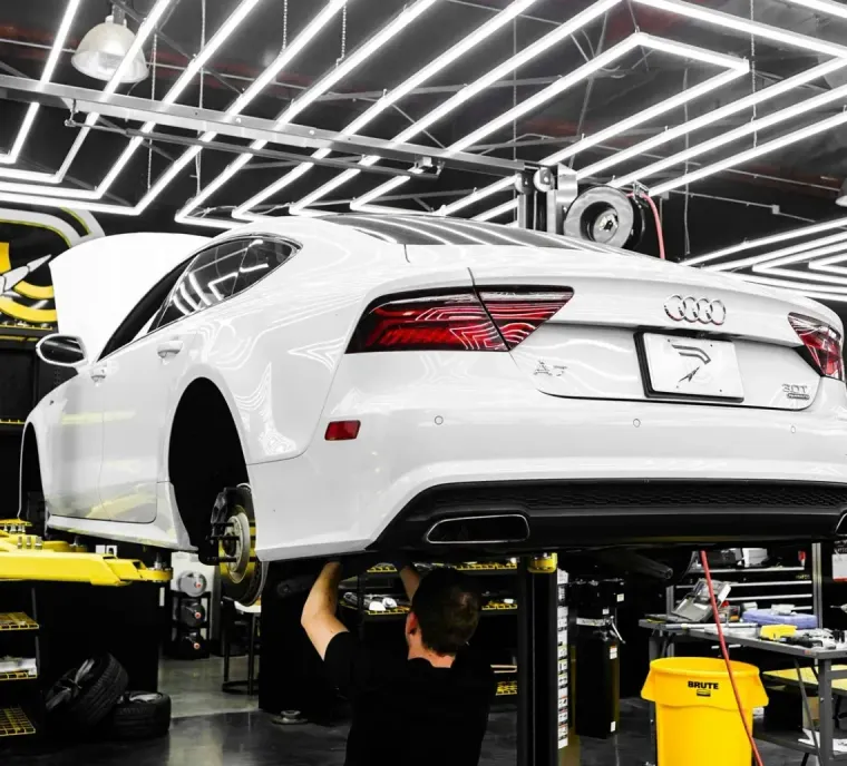 White Audi sedan on a lift in a well-lit auto shop, mechanic working underneath.