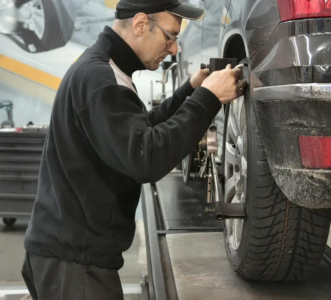 A mechanic wearing a black cap and sweater performs a wheel alignment on a dark vehicle in an auto repair shop.