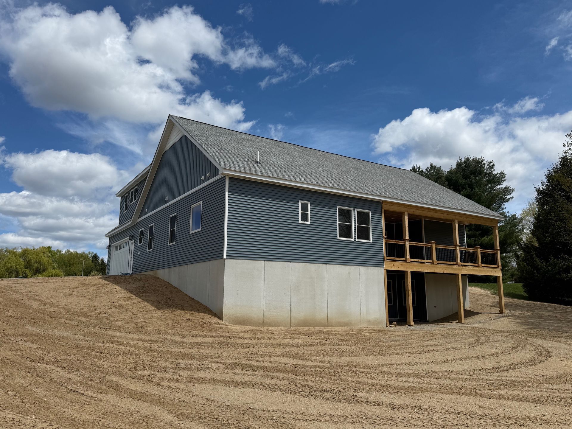 Gray house with a wooden deck on a sloped dirt lot under a partly cloudy blue sky