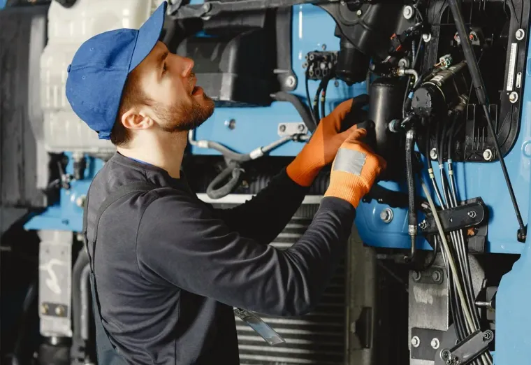 Mechanic in blue cap and orange gloves working on a vehicle engine in a garage
