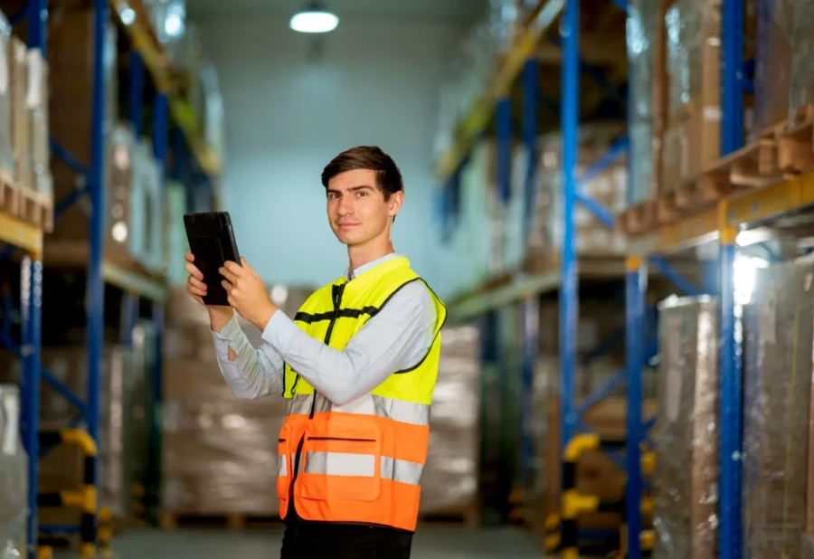 Warehouse worker in a neon vest using a tablet between tall storage racks.
