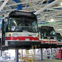 Two buses lifted on maintenance hoists inside a garage, with red-and-white fronts visible.