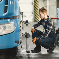 Mechanic in blue and plaid crouches beside a blue truck, inspecting the wheel area in a garage.