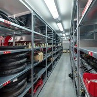A narrow warehouse aisle lined with metal shelves holding black parts and red bins under bright fluorescent lights