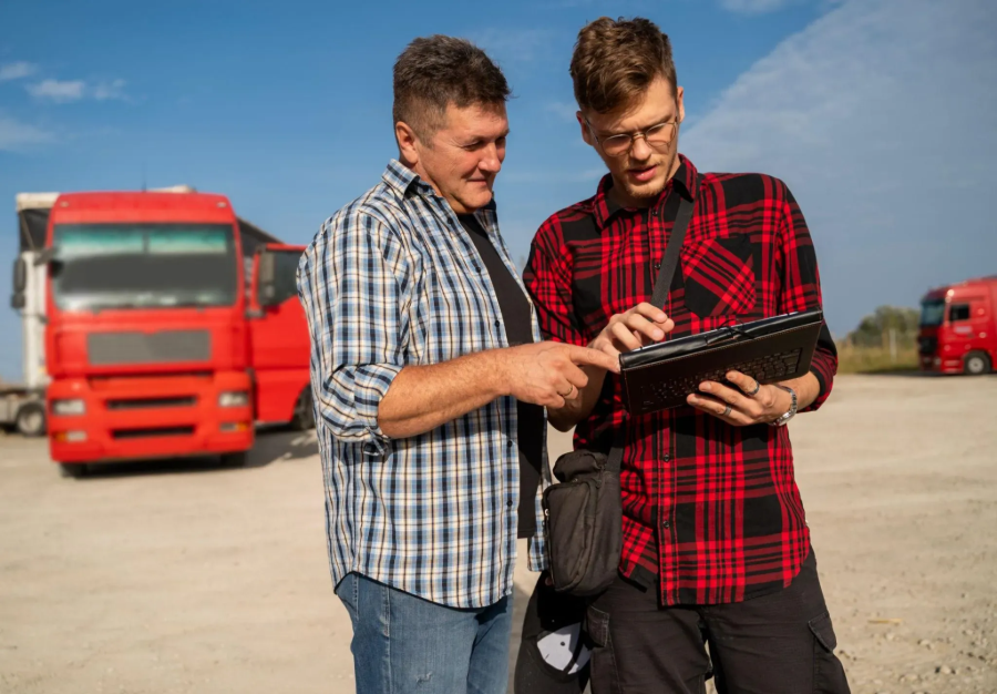Two men in plaid shirts reviewing a clipboard in a truck lot with red trucks in the background