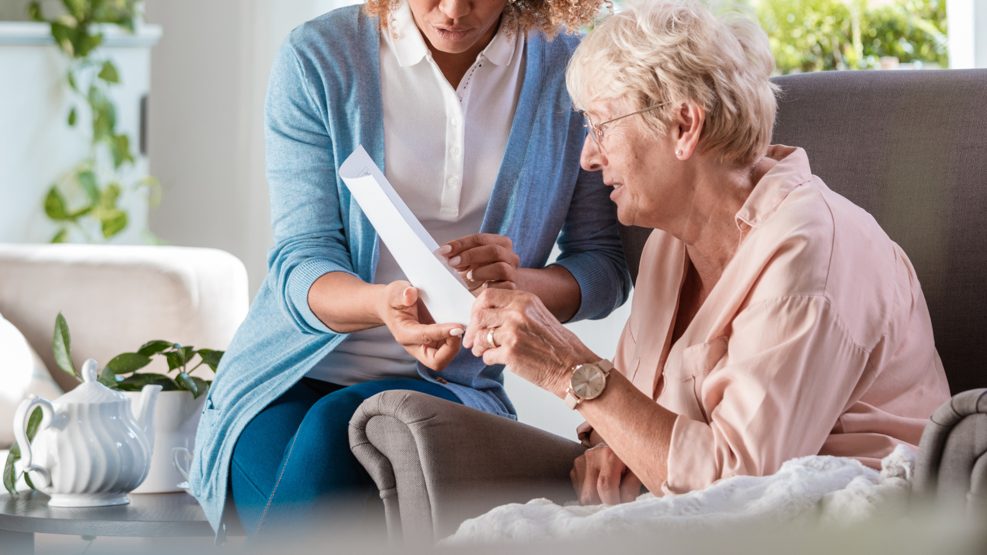 An elderly woman is sitting in a chair while a woman holds her hand and looks at a piece of paper.