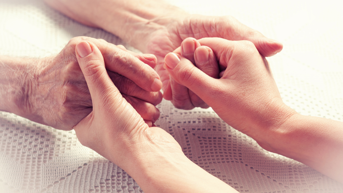 A woman is holding the hand of an elderly man while sitting in a chair.