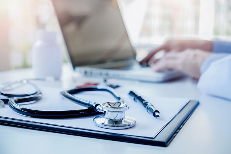 A stethoscope is sitting on top of a clipboard next to a laptop.