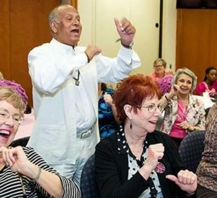 Man in white outfit leads a group of people dancing in a room. People wear pink and smile.