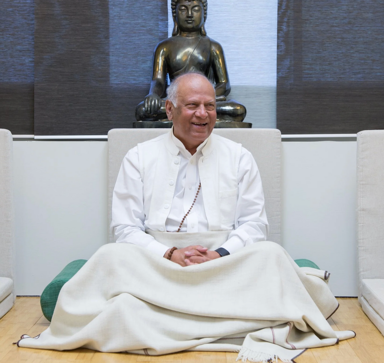 Man sitting cross-legged, smiling, with a Buddha statue behind him. He's in white clothing and a light blanket.