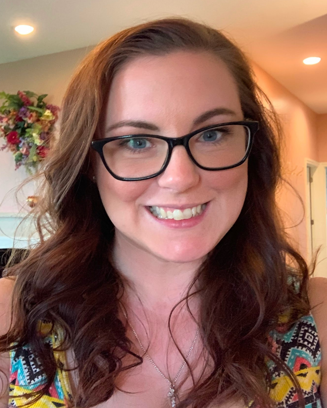 Smiling person with glasses in a colorful top, indoors with warm lighting and a blurred floral background